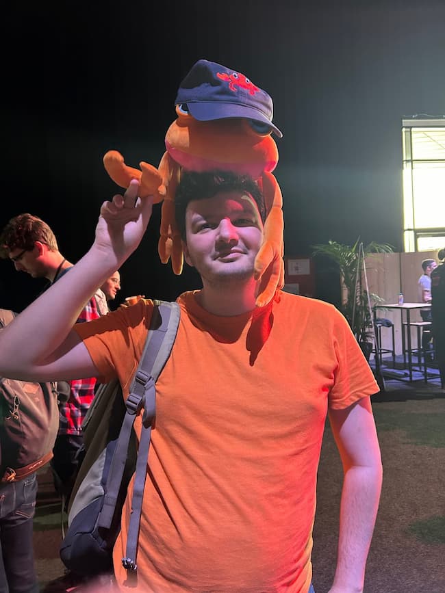 A man in a bright orange shirt wearing the Nova plush as a hat, with a real hat on top of that.